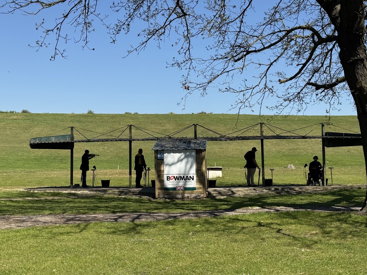Shooters on the covered DTL stands at the North of England Clay Target Centre on a sunny day, with the grass bank behind